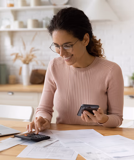 Fröhliche Frau sitzt am Küchentisch mit mehreren Unterlagen, einem Laptop sowie einem Taschenrechner vor sich.